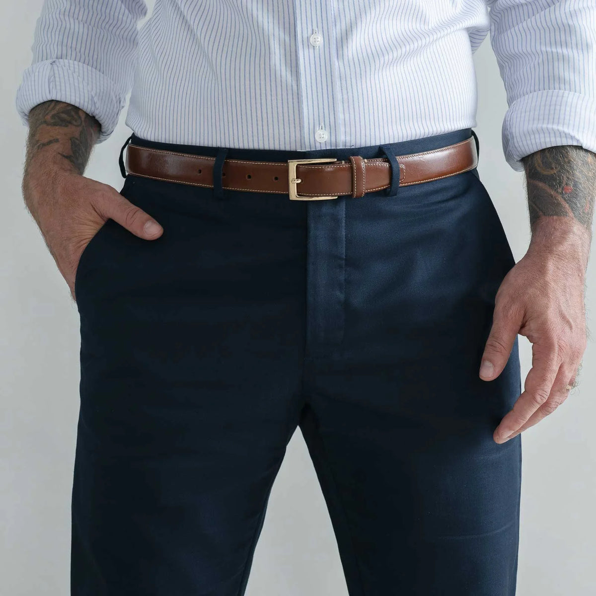 A man wearing navy blue dress pants, a brown leather belt with a gold buckle, and a white and blue striped button-down shirt, shown from the chest down.