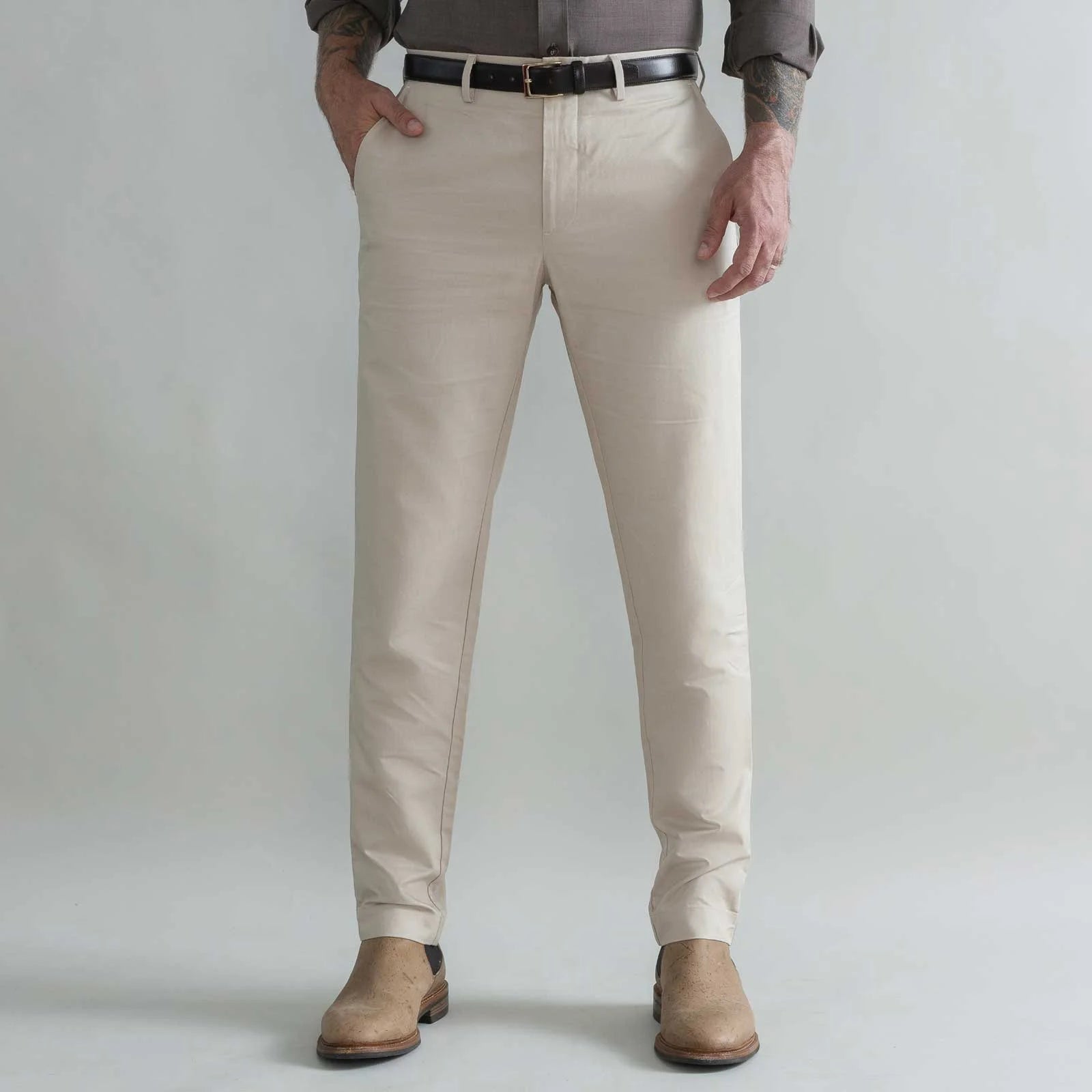 A man wearing khaki slim-fit chinos with a black belt, a brown shirt, and tan boots, standing against a light gray studio background.
