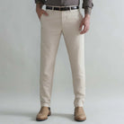 A man wearing khaki slim-fit chinos with a black belt, a brown shirt, and tan boots, standing against a light gray studio background.