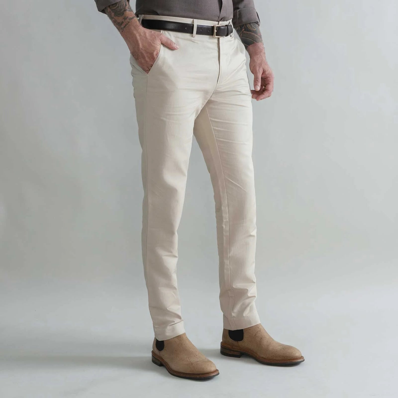 A man wearing slim-fit khaki trousers, styled with a black belt, a gray shirt, and tan suede Chelsea boots, shown from the waist down against a light gray studio background.