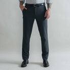 A man wearing dark charcoal gray wool trousers, a black leather belt, a light gray shirt, and black leather loafers, standing against a light gray studio background.