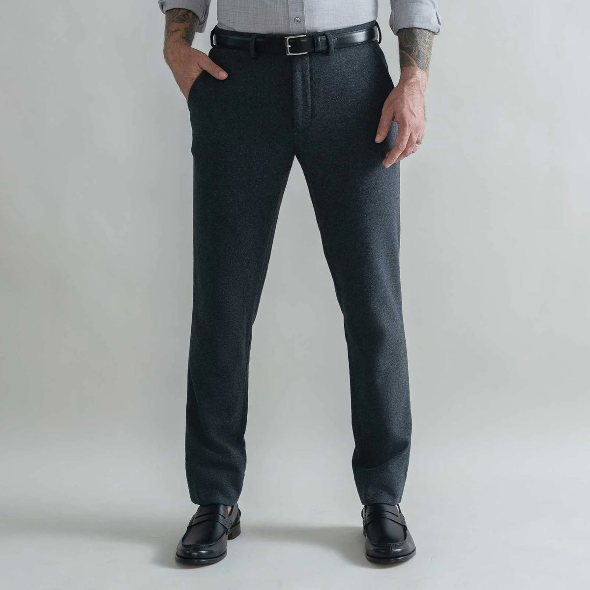 A man wearing dark charcoal gray wool trousers, a black leather belt, a light gray shirt, and black leather loafers, standing against a light gray studio background.
