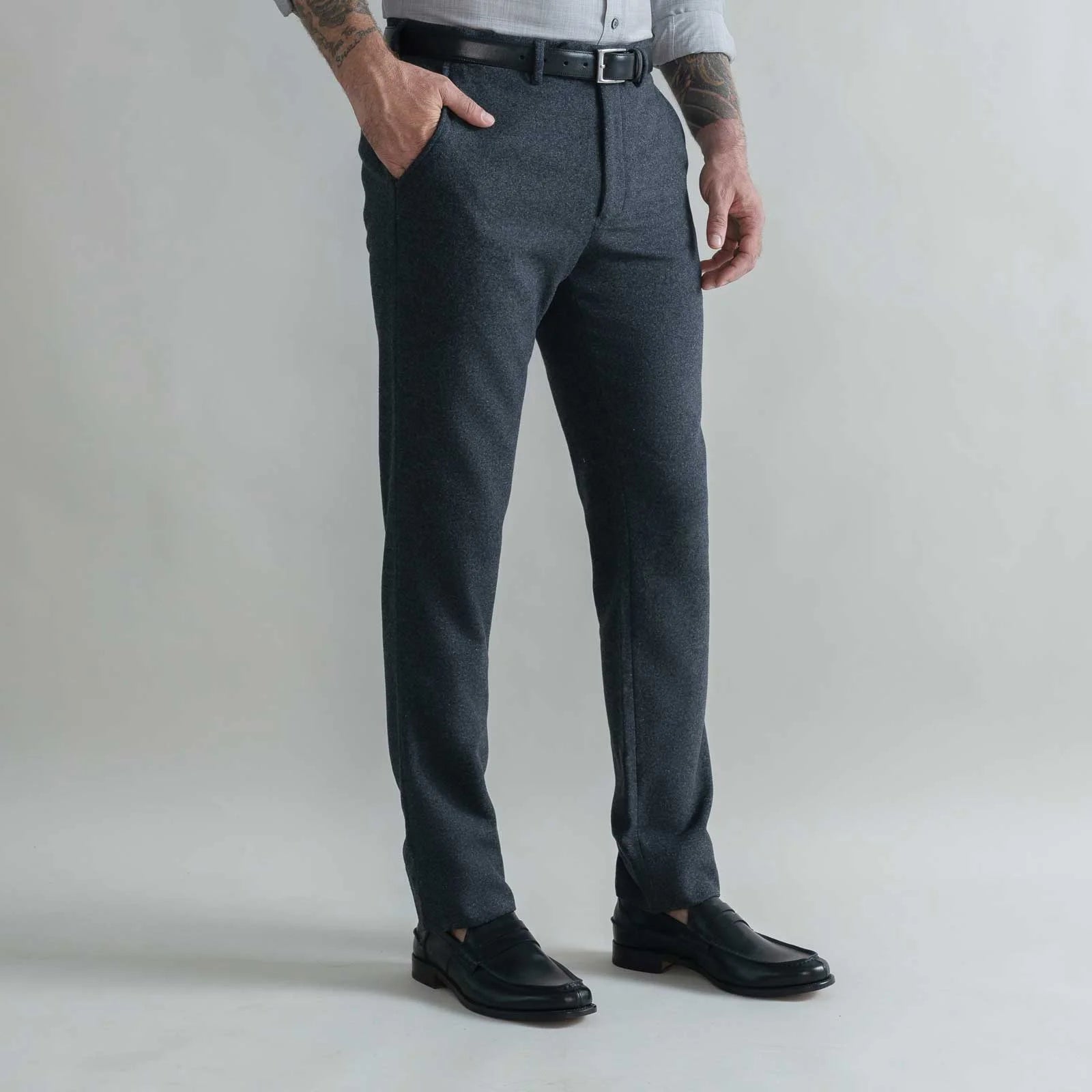 A man wearing dark gray heathered wool trousers, a black belt, a light gray shirt, and black leather loafers, standing against a light gray studio background.