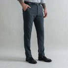 A man wearing dark gray heathered wool trousers, a black belt, a light gray shirt, and black leather loafers, standing against a light gray studio background.