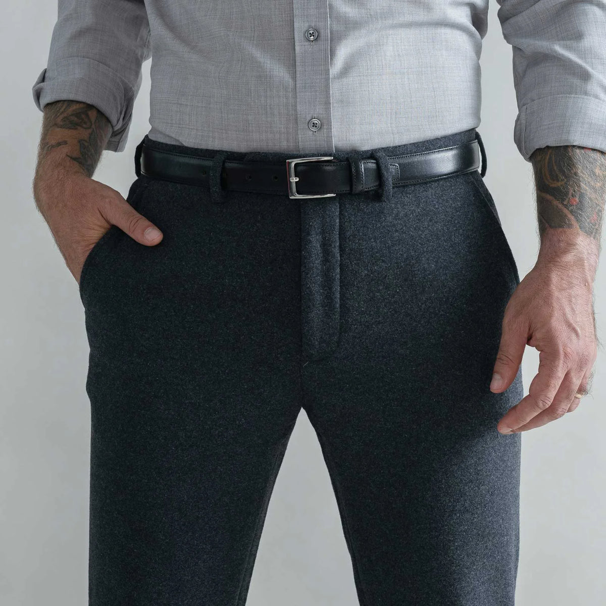 A man wearing dark charcoal gray wool trousers, a light gray button-down shirt, and a black leather belt with a silver buckle, shown from the waist down against a light gray background.