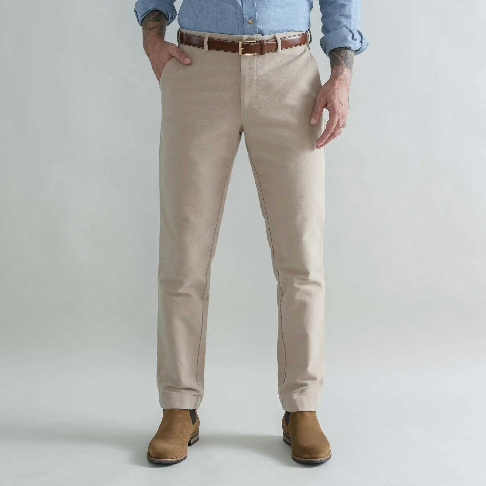 A man wearing tan moleskin chino pants with a brown belt, a light blue button-down shirt, and brown boots, standing against a light gray studio background.