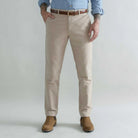 A man wearing tan moleskin chino pants with a brown belt, a light blue button-down shirt, and brown boots, standing against a light gray studio background.
