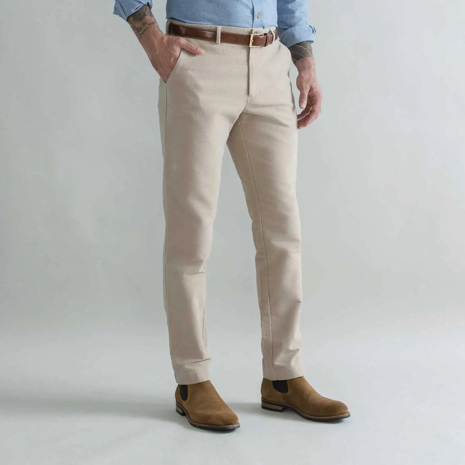 A man wearing tan moleskin chinos with a brown belt, a light blue shirt, and brown suede boots, standing against a light gray studio background.