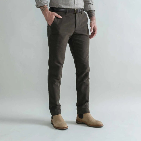 A man wearing slim-fit dark brown moleskin trousers, a gray shirt, and tan Chelsea boots, standing against a light gray studio background.