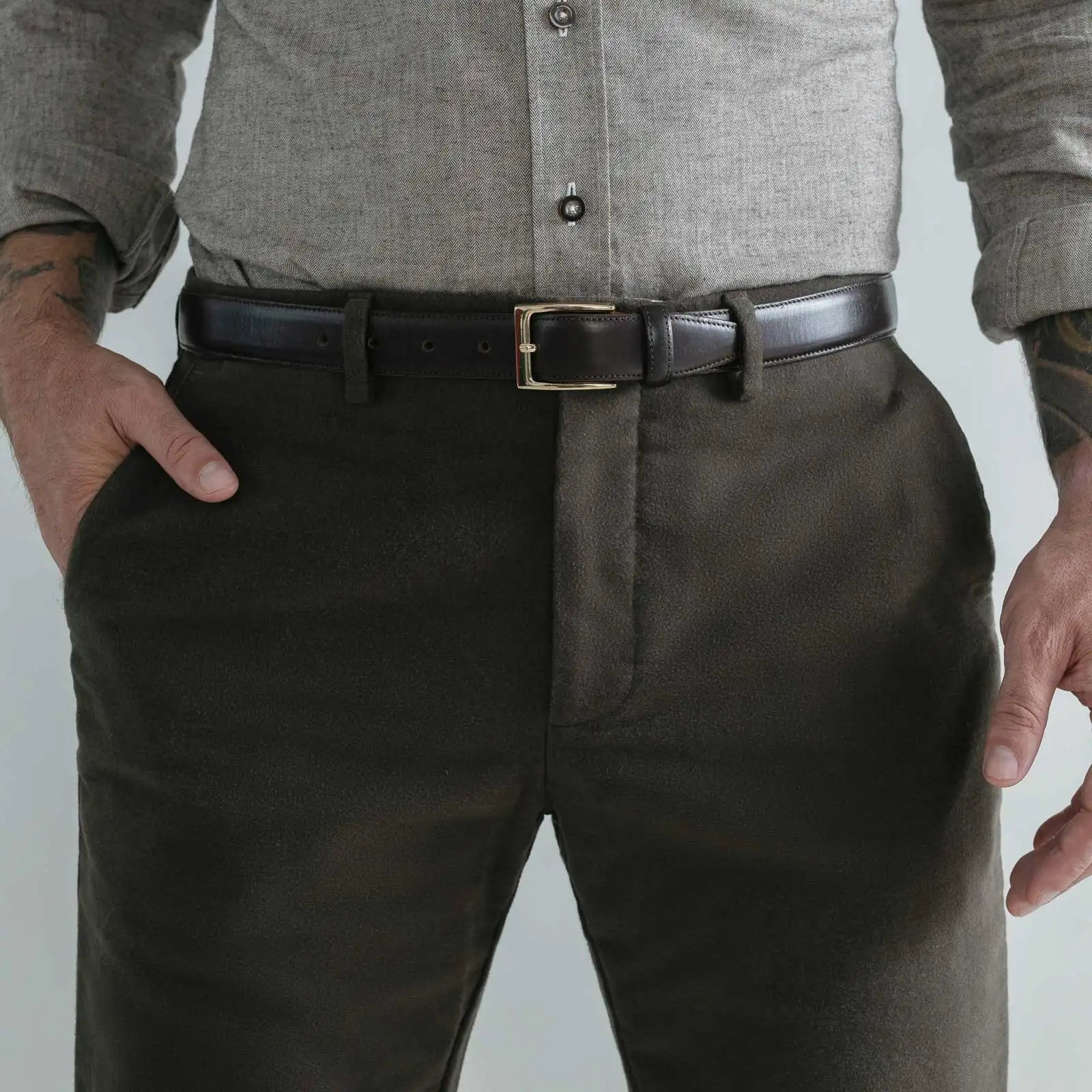 A close-up of a man wearing dark brown moleskin trousers, a black leather belt with a gold buckle, a light gray button-down shirt, and a dark shirt underneath.