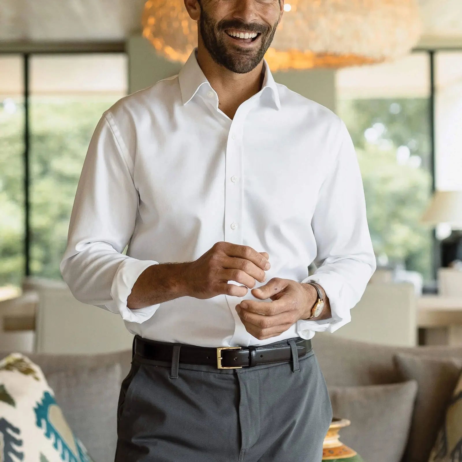 A man wearing a crisp white long-sleeve button-down dress shirt, dark gray trousers, and a black belt with a gold buckle, standing in a modern living room setting.