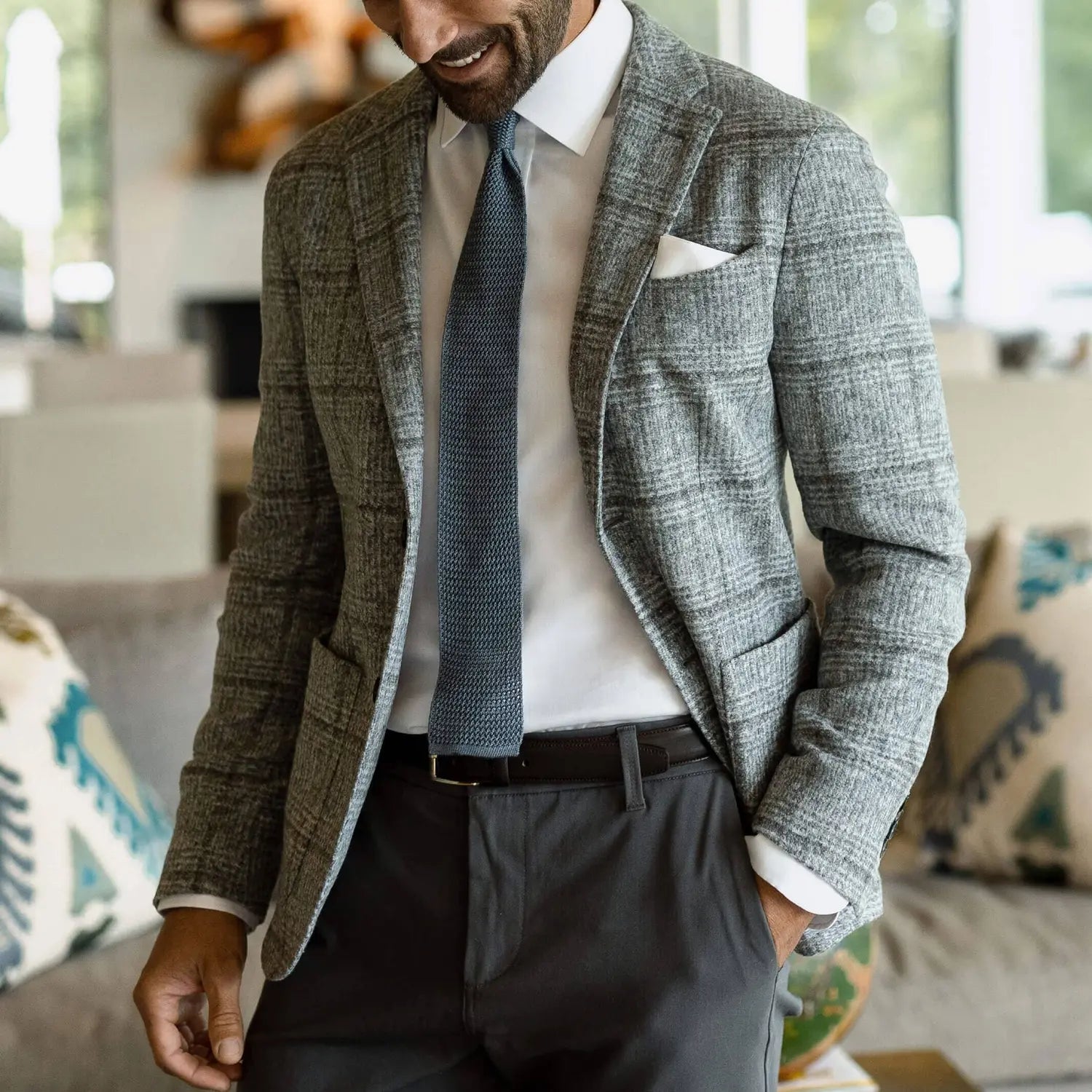 A man wearing a gray and white plaid blazer, white dress shirt, textured blue tie, and dark gray trousers, standing in a modern living room setting.