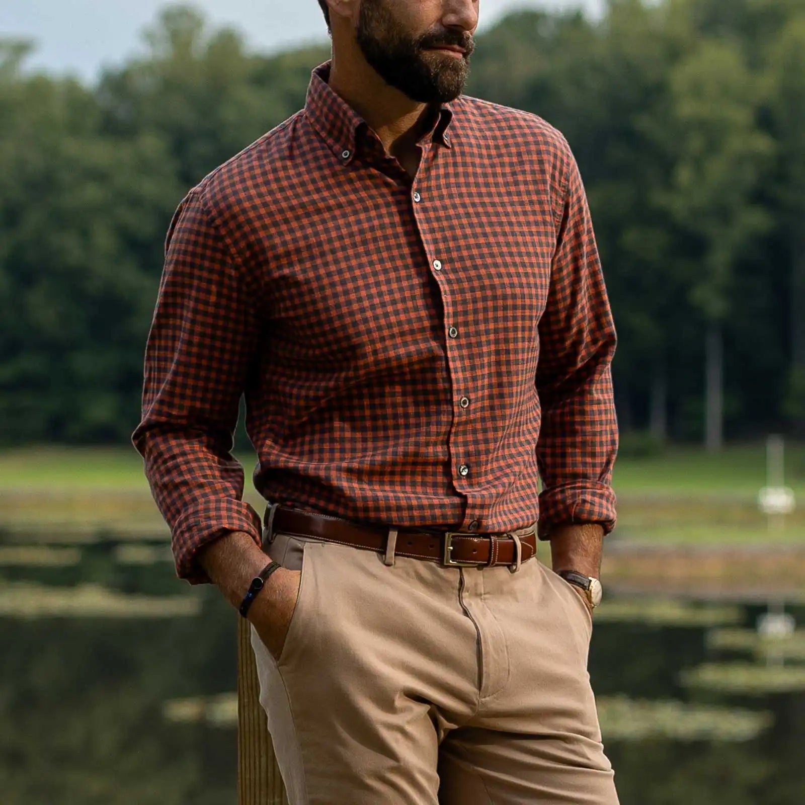 A man wearing a rust-orange and brown checkered long-sleeve button-down shirt, khaki pants, and a brown belt, standing outdoors near a lake with trees in the background.