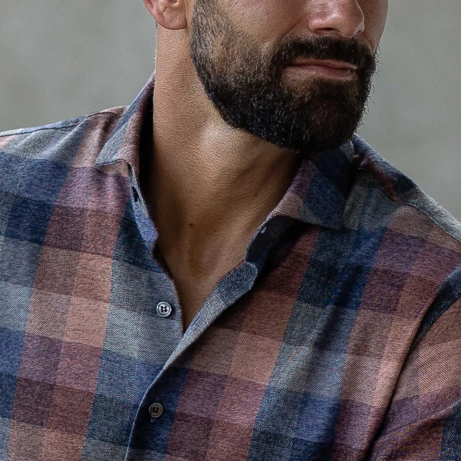 A close up of a plaid shirt in muted blues and orange tones on a man with a beard looking off to the side, with his eyes cut off just above the nose.
