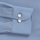 Close-up of a light blue dress shirt cuff with two white mother of pearl buttons on a matching blue fabric background.
