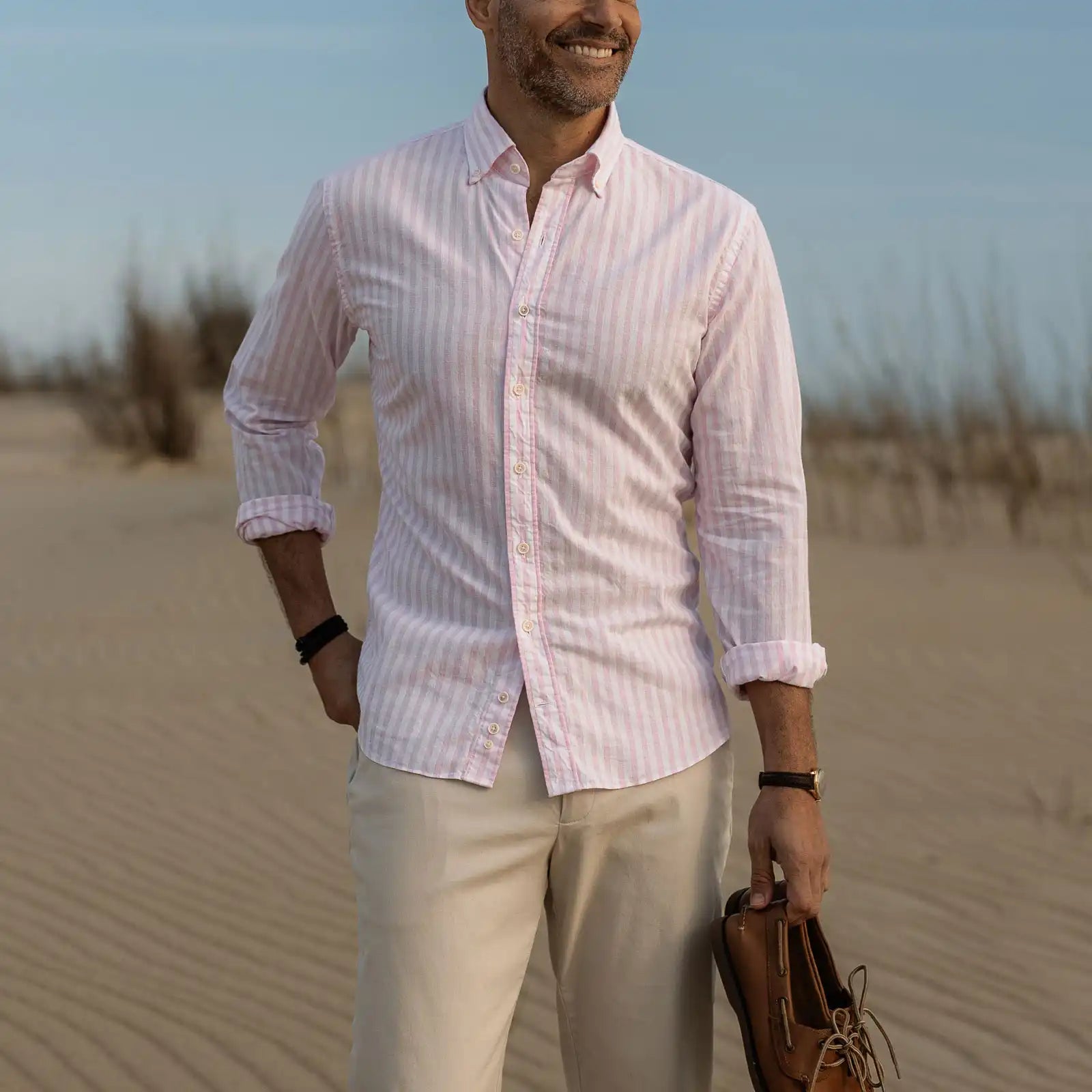 Man in a pink striped shirt and beige pants standing on sand with a clear sky.