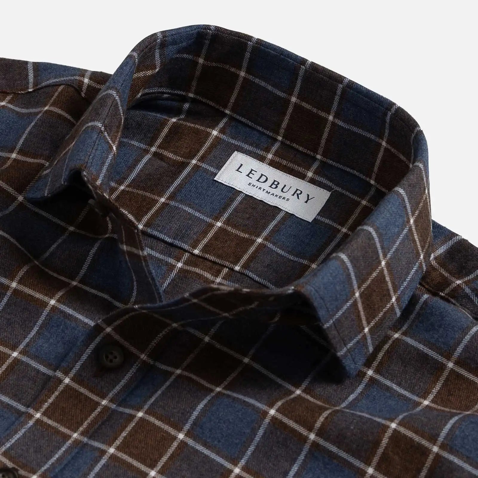 Close-up of a plaid button-down shirt collar with a visible LEDBURY brand label. The fabric is a mix of dark brown, slate blue, and thin white grid lines.