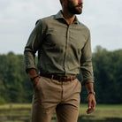 A man wearing a green checkered long-sleeve button-down shirt, khaki pants, and a brown belt, standing outdoors in a grassy field with trees in the background.