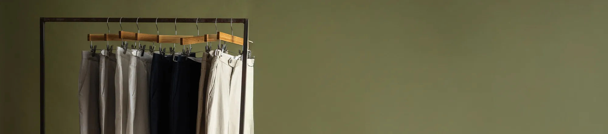 Clothing rack with several pairs of Ledbury pants in different colors hanging on wooden hangers in front of an olive green studio background.