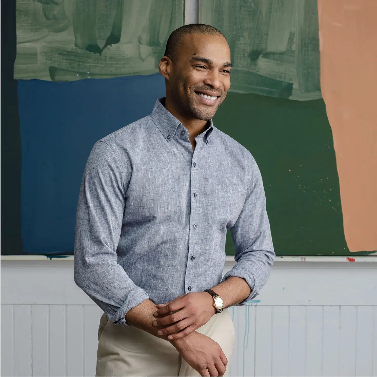 Model wearing a men's pale navy chambray button down shirt. He is standing in an artist's studio in front of a painting. 