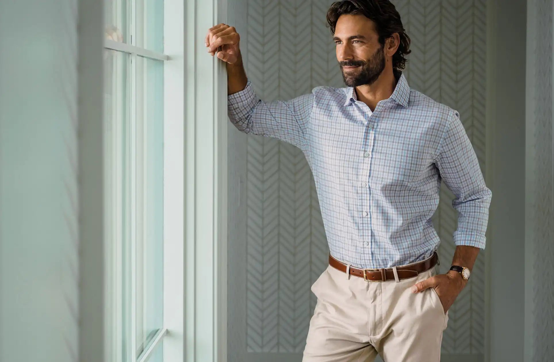 Man wearing a checkered shirt and beige pants standing by a window.