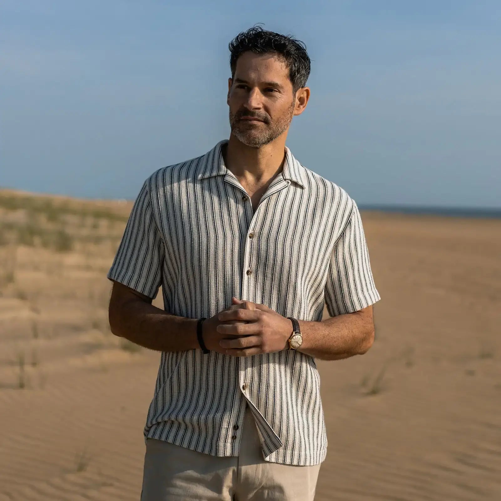 Man standing in on the beach wearing a striped short sleeve camp collar shirt, shorts and watch.