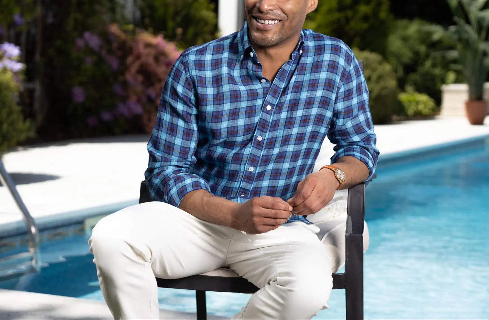 Male model sitting in a chair next to a pool wearing a bright blue and dark blue plaid linen shirt 