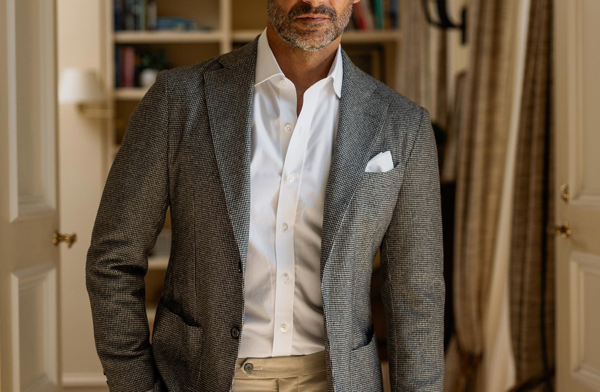 Man wearing a gray blazer over a white shirt with a beige pocket square, standing indoors.