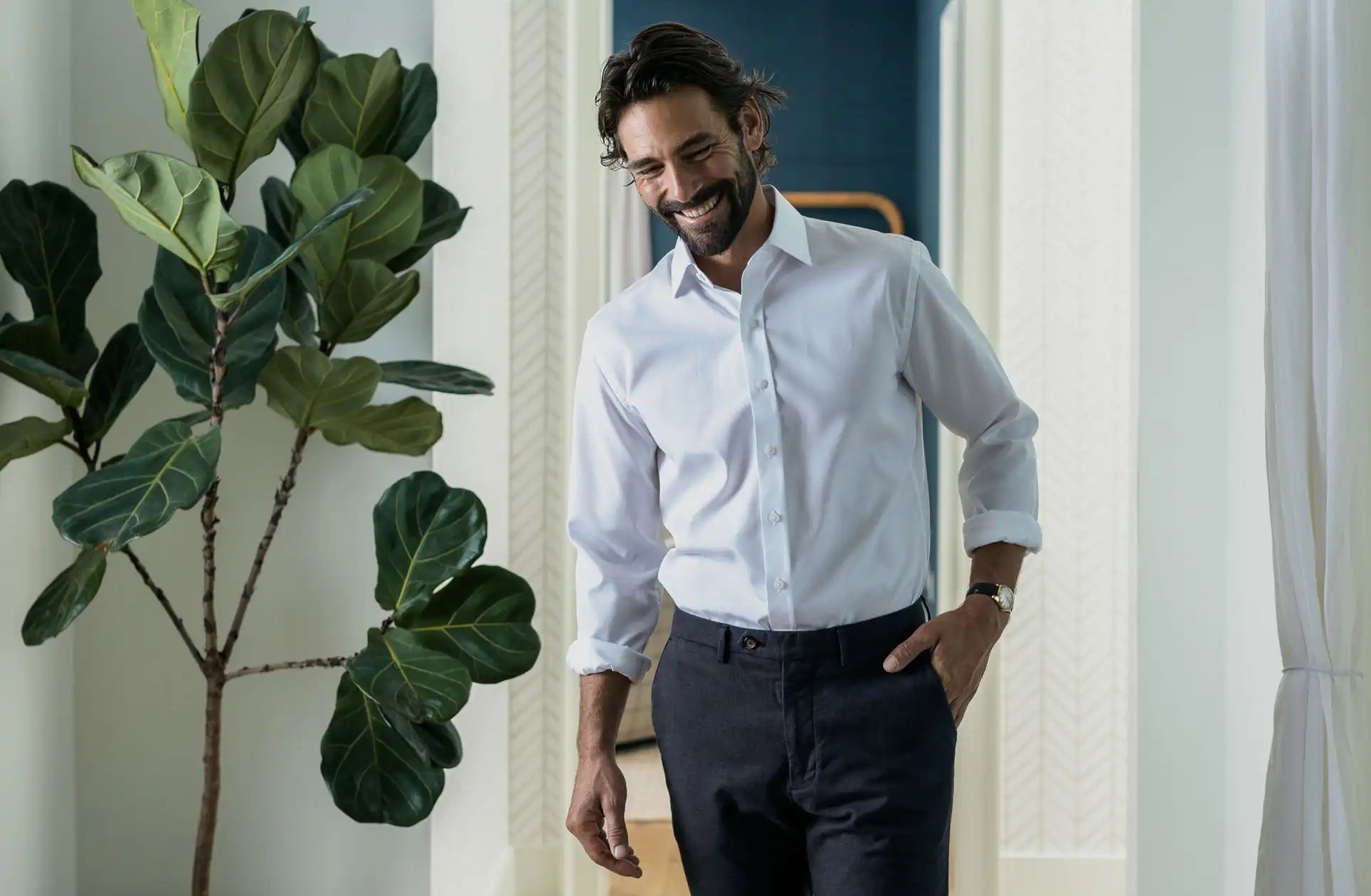 Man wearing a white dress shirt and dark pants standing indoors with a plant to his left.