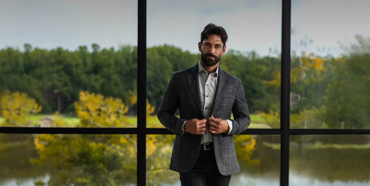 A man wearing a gray plaid blazer over a white shirt stands in front of large black-framed windows with a blurred outdoor landscape of trees and water in the background.