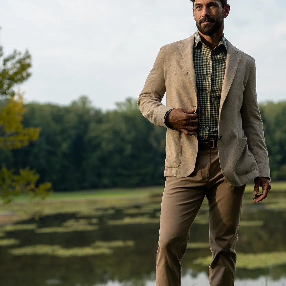 A man wearing a beige blazer, tan trousers, a green and black checkered shirt, and a brown belt stands outdoors near a lake with trees in the background.
