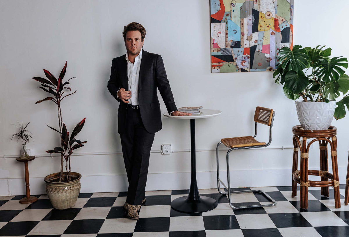 Man in a suit standing next to a small table and chair in a room with checkered floor and plants.