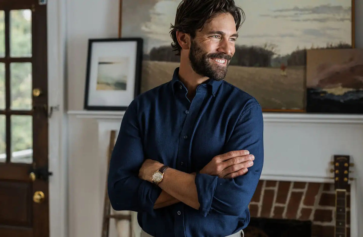 Male model wearing a Ledbury dark navy denim button down dress shirt. He is standing in front of a fireplace. 