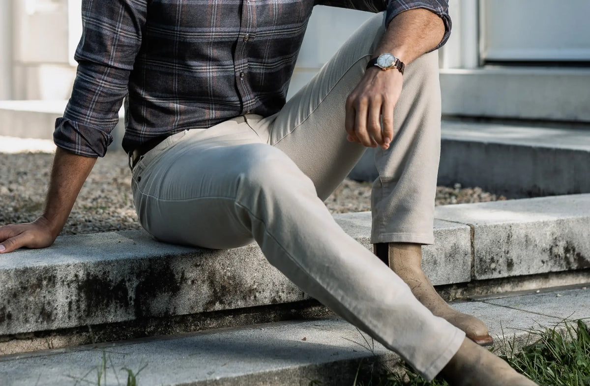 A man sitting on concrete steps outdoors, wearing light beige chinos, a dark gray plaid button-down shirt, and brown boots.