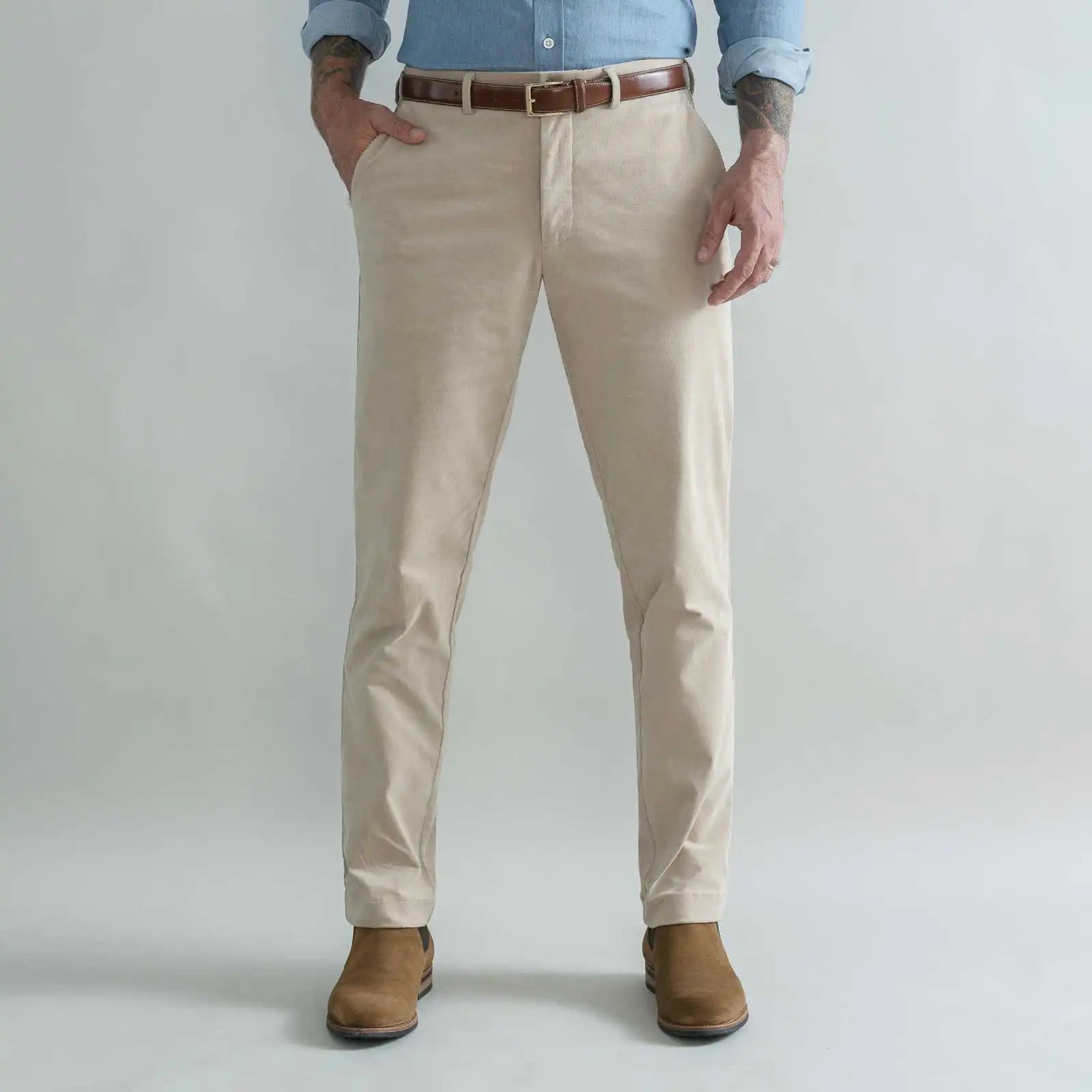 A man wearing tan corduroy chino pants with a brown belt, a light blue button-down shirt, and brown boots, standing against a light gray studio background.
