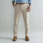 A man wearing tan corduroy chino pants with a brown belt, a light blue button-down shirt, and brown boots, standing against a light gray studio background.