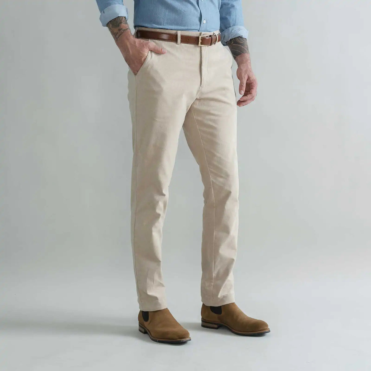 A man wearing tan slim-fit corduroy chinos with a brown belt, a light blue button-down shirt, and brown suede boots, standing against a light gray studio background.