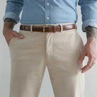 A close-up of a person wearing tan corduroy chinos with a brown leather belt and a light blue button-down shirt, cropped from the chest down against a light gray background.