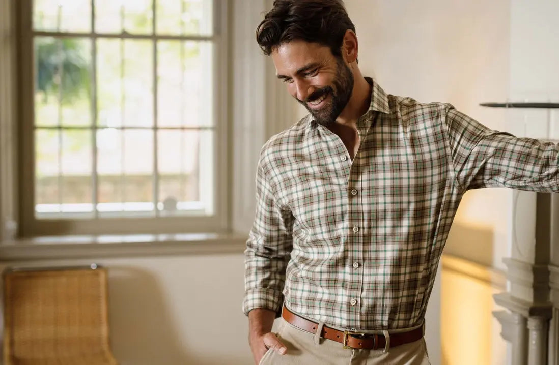 A smiling man wearing a long-sleeve plaid button-down shirt in beige, brown, and green tones, paired with khaki pants and a brown belt, standing indoors near a window.