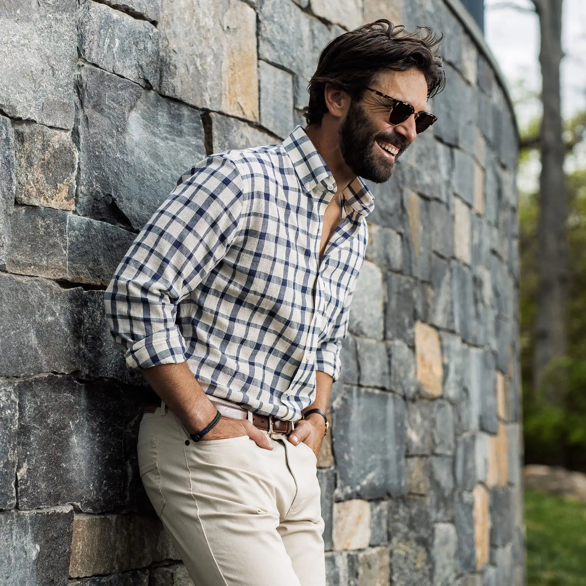 Model leaning up against a stone wall in an outdoor setting. He is wearing a Ledbury navy and tan plaid breathable cotton button-down casual shirt and tan chinos. Clicking this banner will lead to the Casual Shirts collection.
