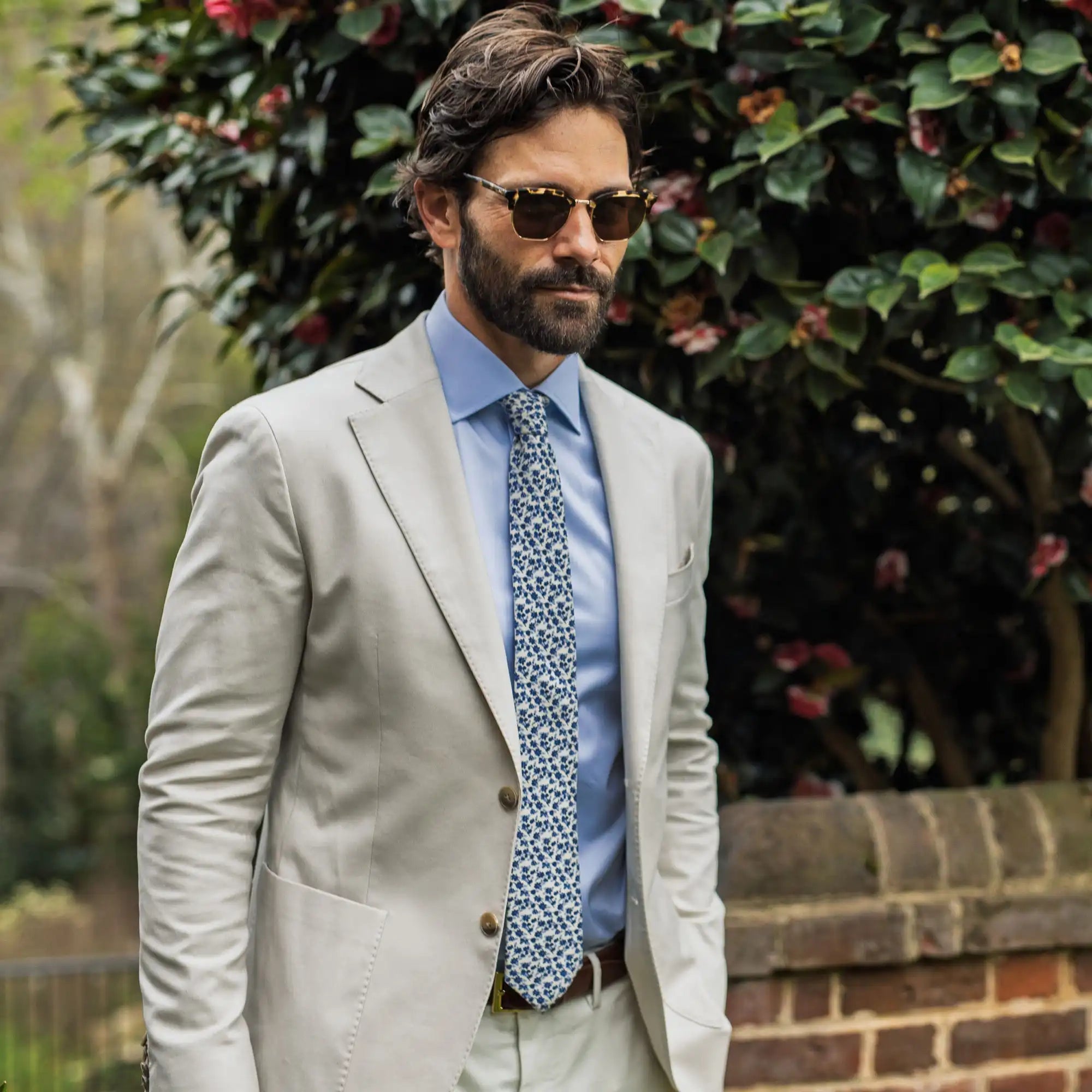 Male model wearing a blue Ledbury dress short, floral tie, and tan sport coat. He is standing outside with sunglasses on. 