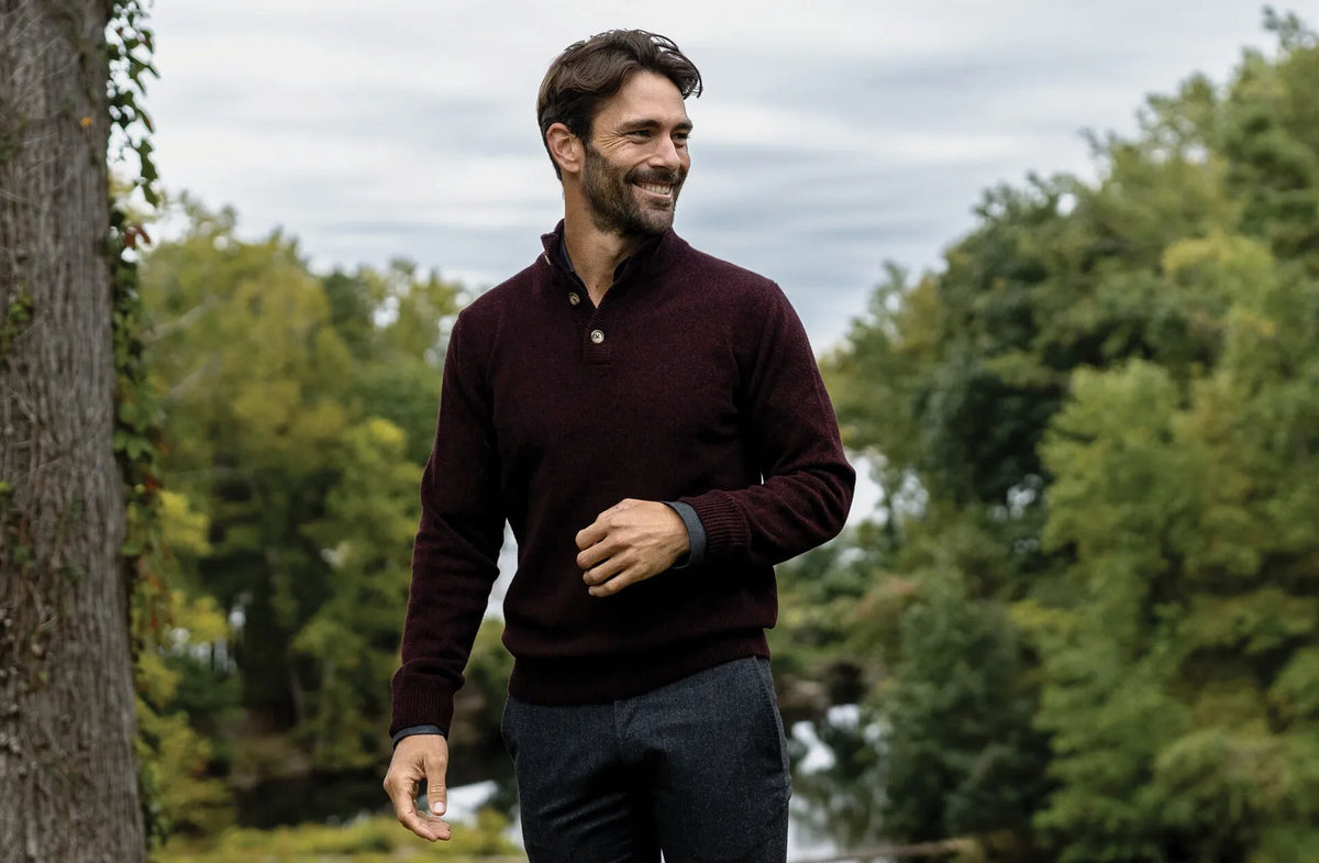 Man wearing a dark red sweater and dark pants standing outdoors with trees in the background